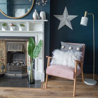 living room with dark blue walls and wooden floor, with a fireplace surrounded by a white mantle, and a pink armchair with fluffy cushions