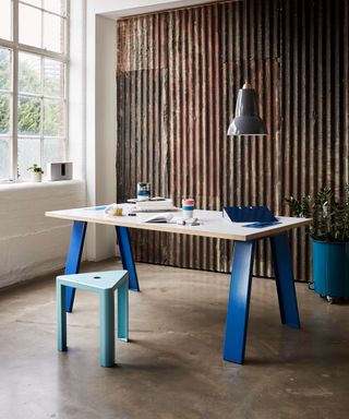 Chunky wooden table with blue metal legs with blue stool in front of industrial metal wall
