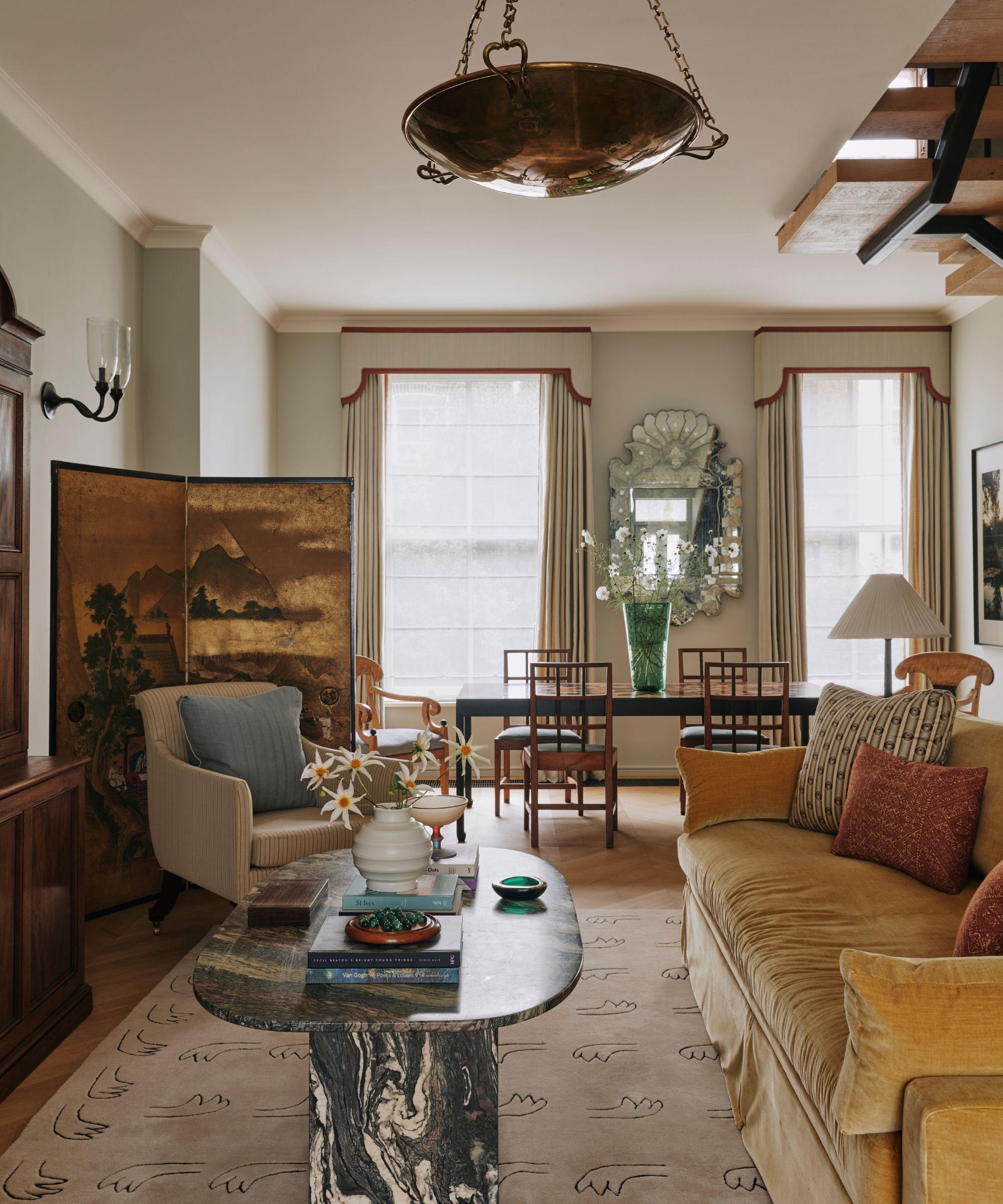 Living room with yellow velvet sofa, marble coffee table, Chinese screen, brass pendant and tall windows with pelmet curtains