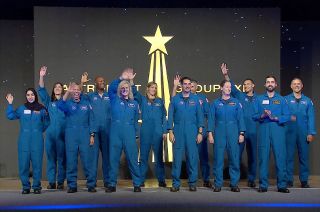 twelve people in blue flight suits stand on a stage and wave to an unseen crowd