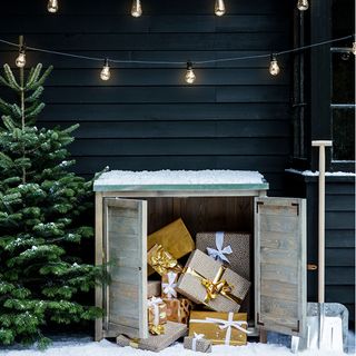 wooden cabinet holding wrapped Christmas presents next to Christmas tree under hanging lights