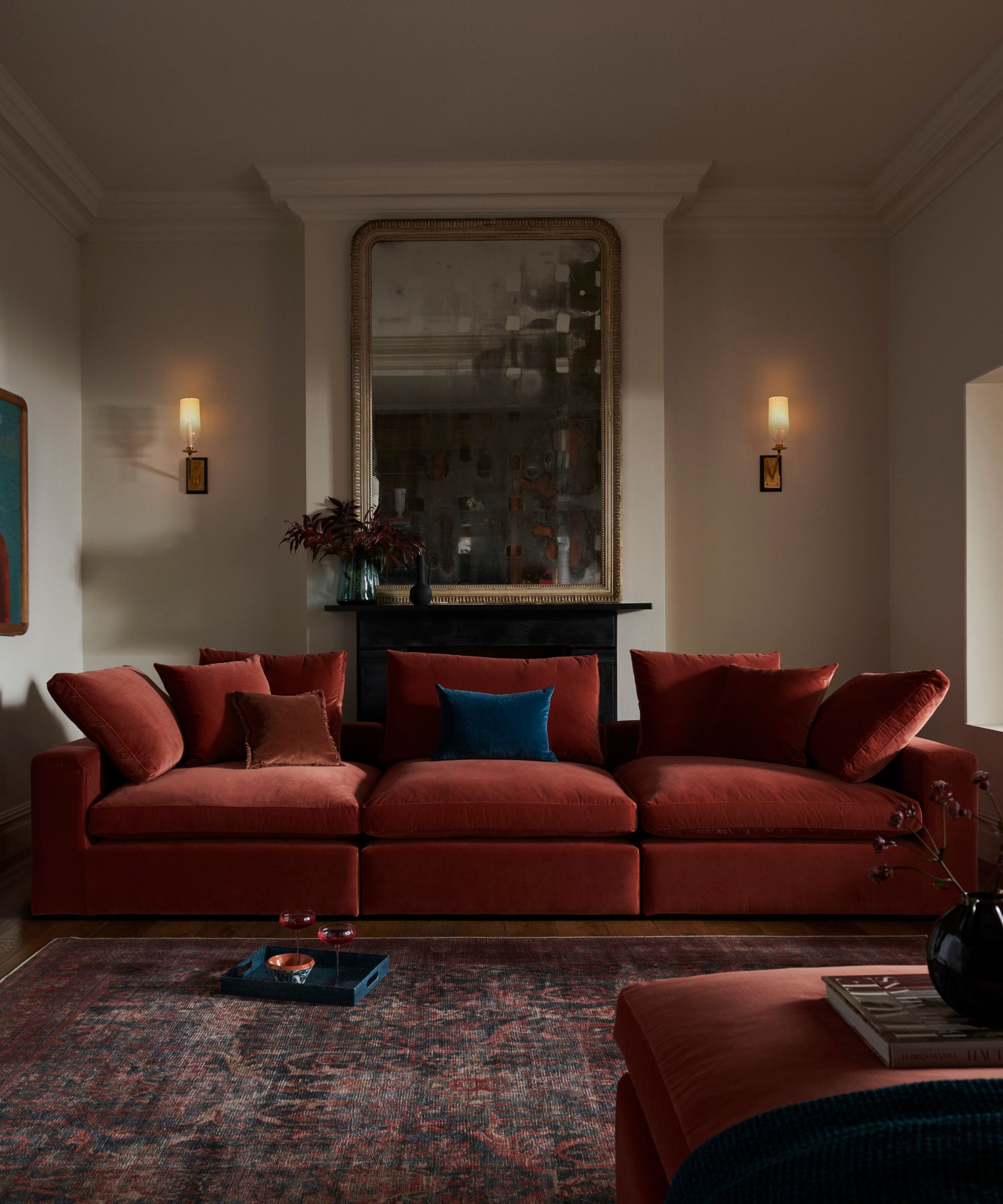 A dramatic living room with a long deep red velvet sofa accented by dark blue and rust-coloured cushions. Two wall sconces cast a soft glow on either side of a large antique mirror above a black fireplace, balancing modern comfort with classic elegance.