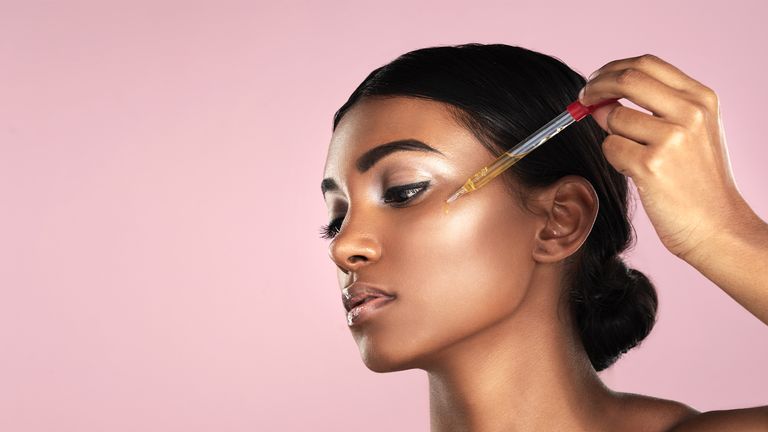 Studio shot of a beautiful young woman applying essential oil to her face with a dropper posing against a pink background