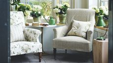 Seating area in sunny conservatory with tiled floors, a floral armchair beside a striped armchair, white flowers, a green watering can on a circular table, and green lamp, surrounded by windows.