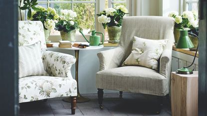 Seating area in sunny conservatory with tiled floors, a floral armchair beside a striped armchair, white flowers, a green watering can on a circular table, and green lamp, surrounded by windows.