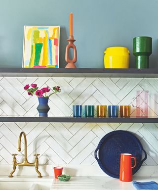 Close up of colorful kitchen shelving with blue painted walls, white tiles, brass tap and marble countertop