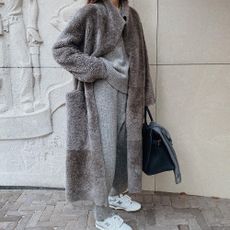 A women stands in front of an ornate concrete wall, the woman wears a grey cashmere sweater with matching trousers, New Balance sneakers and a floor length fleece coat.