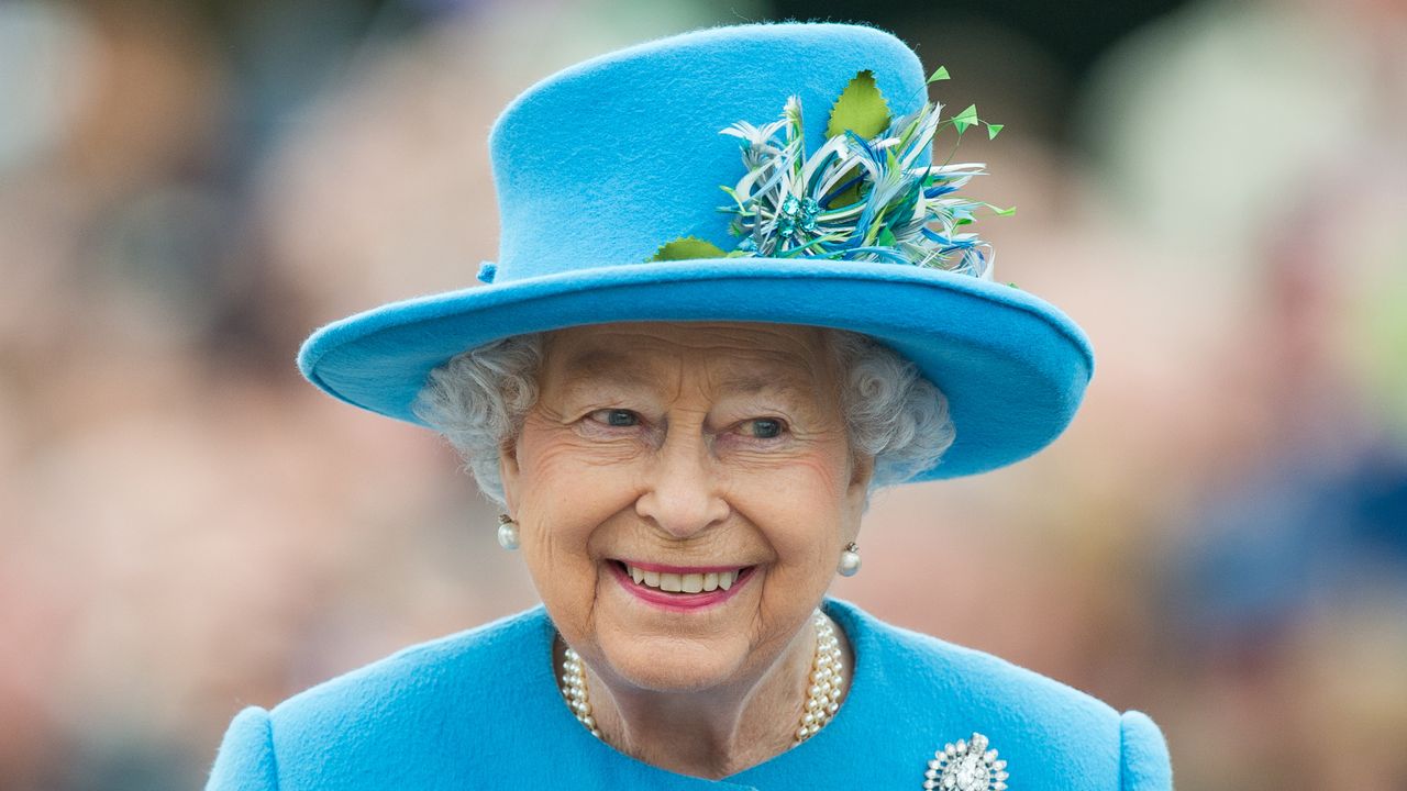 Queen Elizabeth wearing a blue hat with flowers on it and a matching blue coat with pearls and smiling