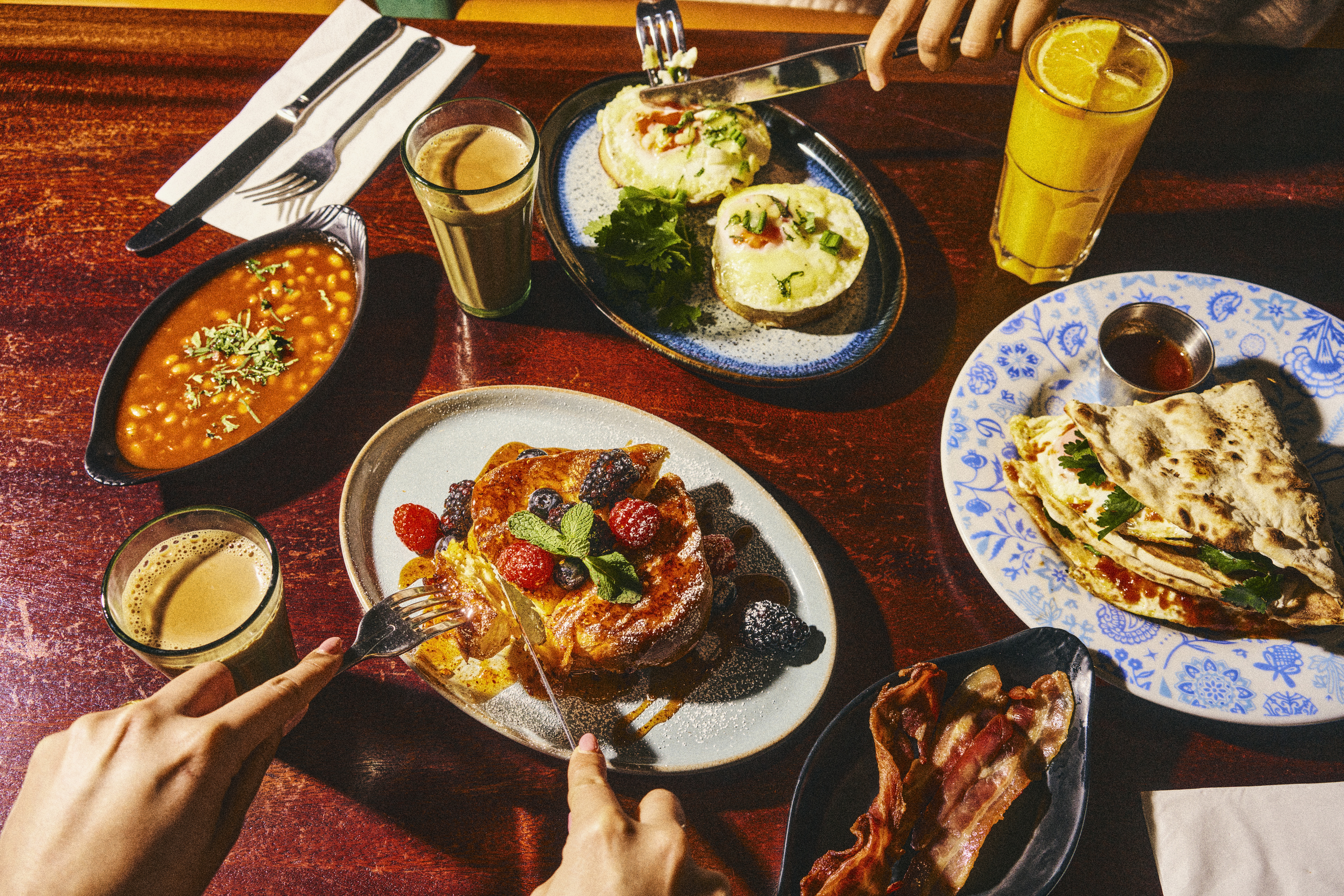 A tantalizing, Indian-British breakfast is served on a wooden table at a restaurant, as shown by a picture shot from above, and showing berry pancakes, baked beans, eggs, and pitas.