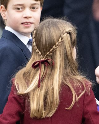 The back of Princess Charlotte's head showing a braided hairstyle tied with a maroon bow and Prince George smiling in the background