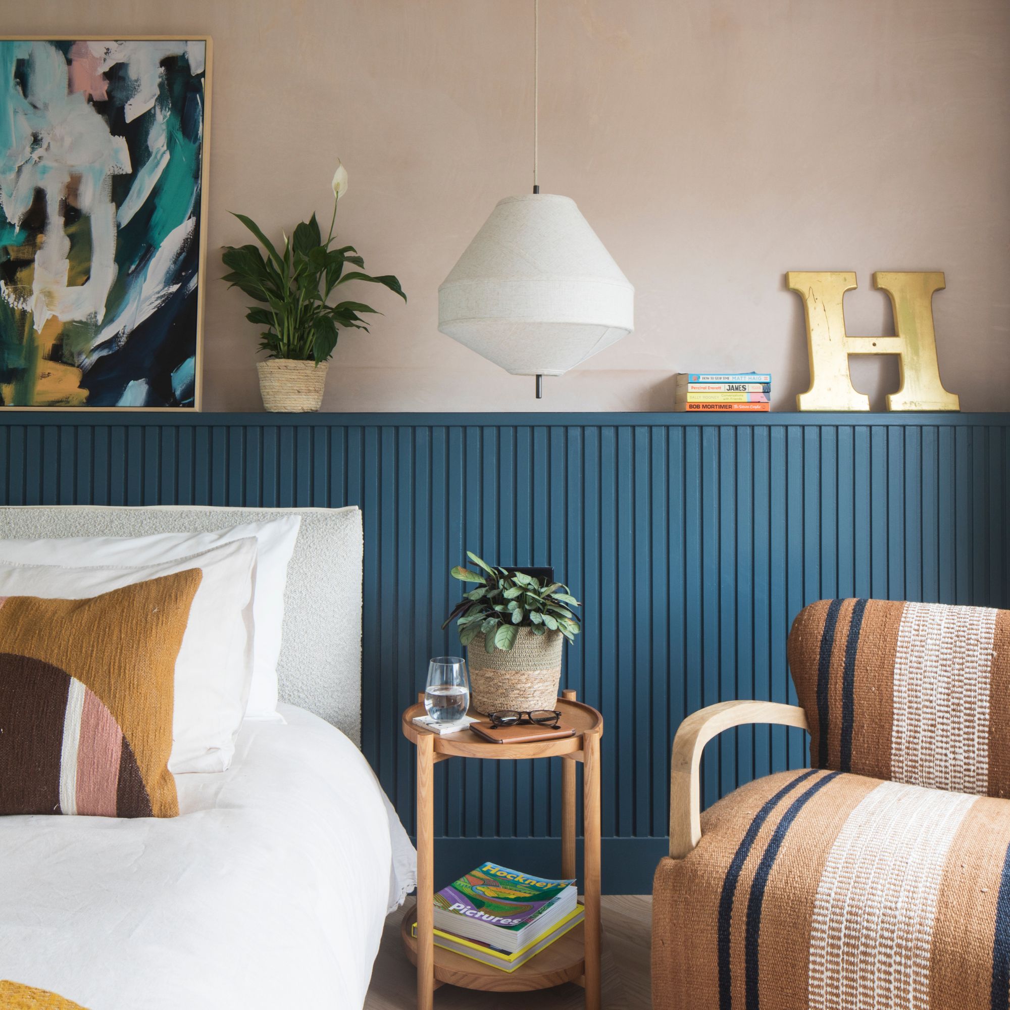 Bedroom corner with pink walls, blue panelling and armchair beside wooden bedside table. Artwork and plant for decor plus a white pendant light.