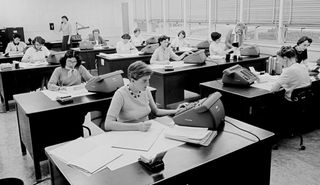 Women sit writing at desks and typing on large calculating machines.