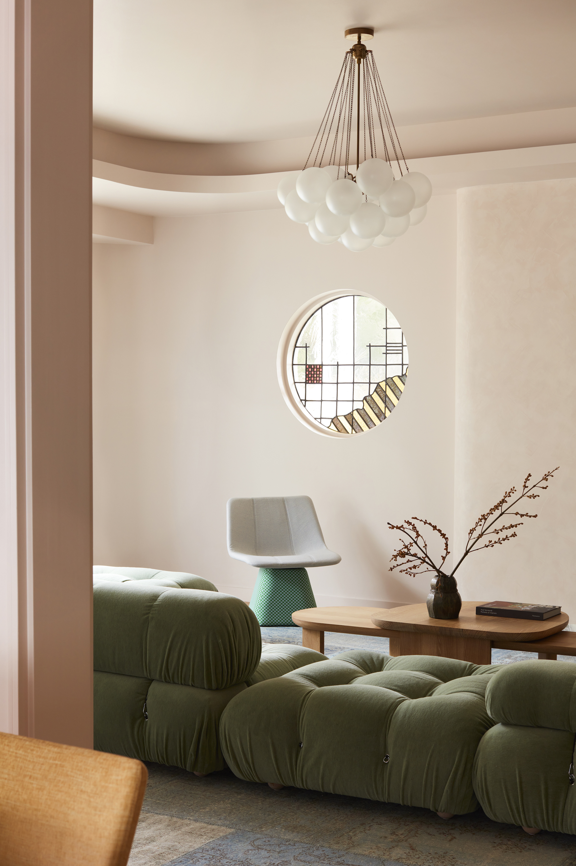 A light, soft pink living space with a round window on the far wall and a modern style chandelier, above a sage green sofa, wooden coffee table and modern armchair.