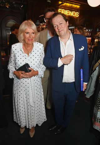 Queen Camilla wearing a white polka dot dress standing next to her son Tom Parker Bowles wearing a blue suit at his book launch party