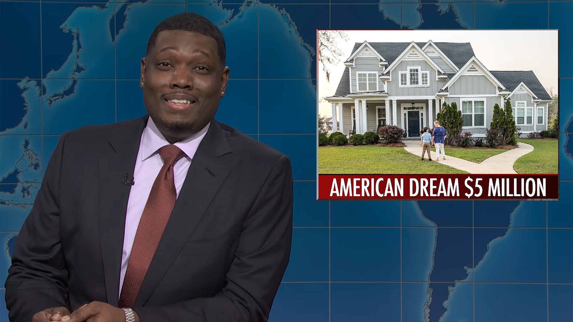 Michael Che in a suit, red tie and pink shirt sits at the Weekend Update desk on SNL. A blue background is behind him. 