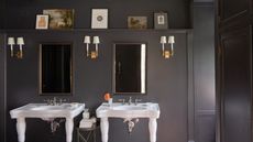 bathroom painted dark gray, double white sinks, mirrors on wall, three walls sconces, shelf with pictures