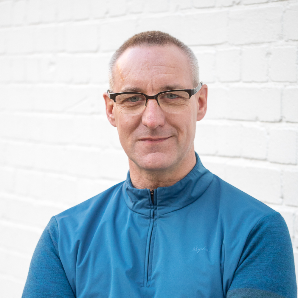 male with short hair, glasses wearing zip up blue top stood against white wall