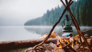 A metal kettle attached to a log structure hangs over a campfire near a lake.