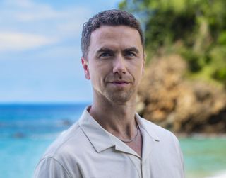 A generic shot of Taylor Fielding (Richard Fleeshman) standing on the beach, smirking at the camera