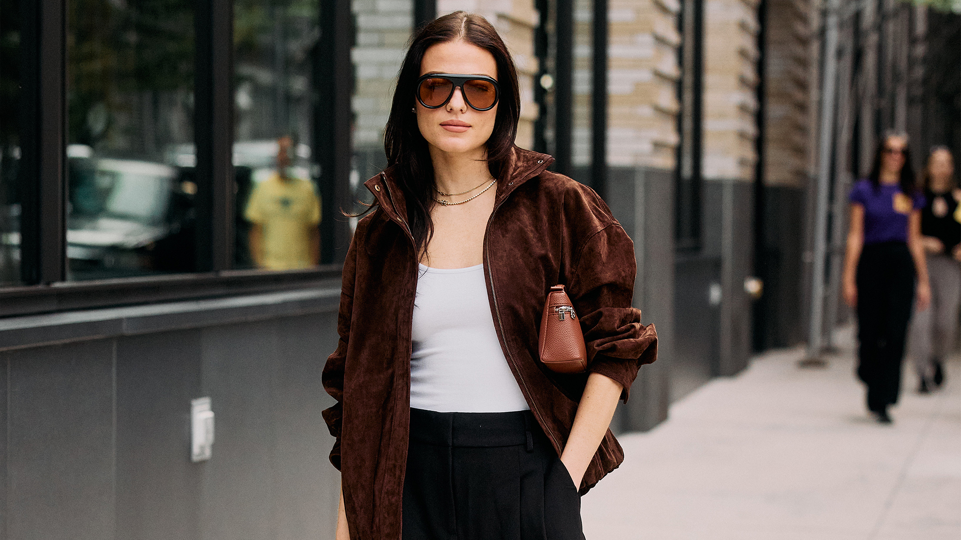 woman wears brown jacket white tank top black pants with a brown handbag tucked under her arm