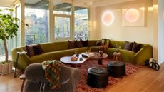 A white living room with windows overlooking a balcony, a large curved green sofa, a brown coffee table, a pair of black pouffes, a green sofa chair with a checked throw