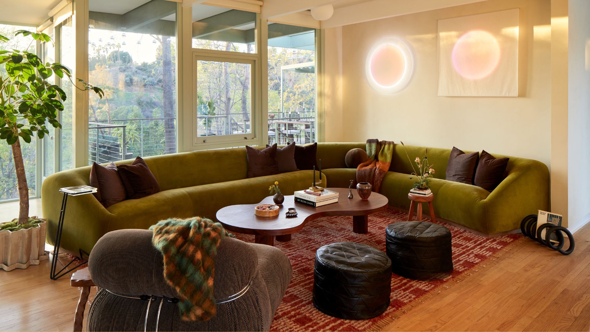 A white living room with windows overlooking a balcony, a large curved green sofa, a brown coffee table, a pair of black pouffes, a green sofa chair with a checked throw