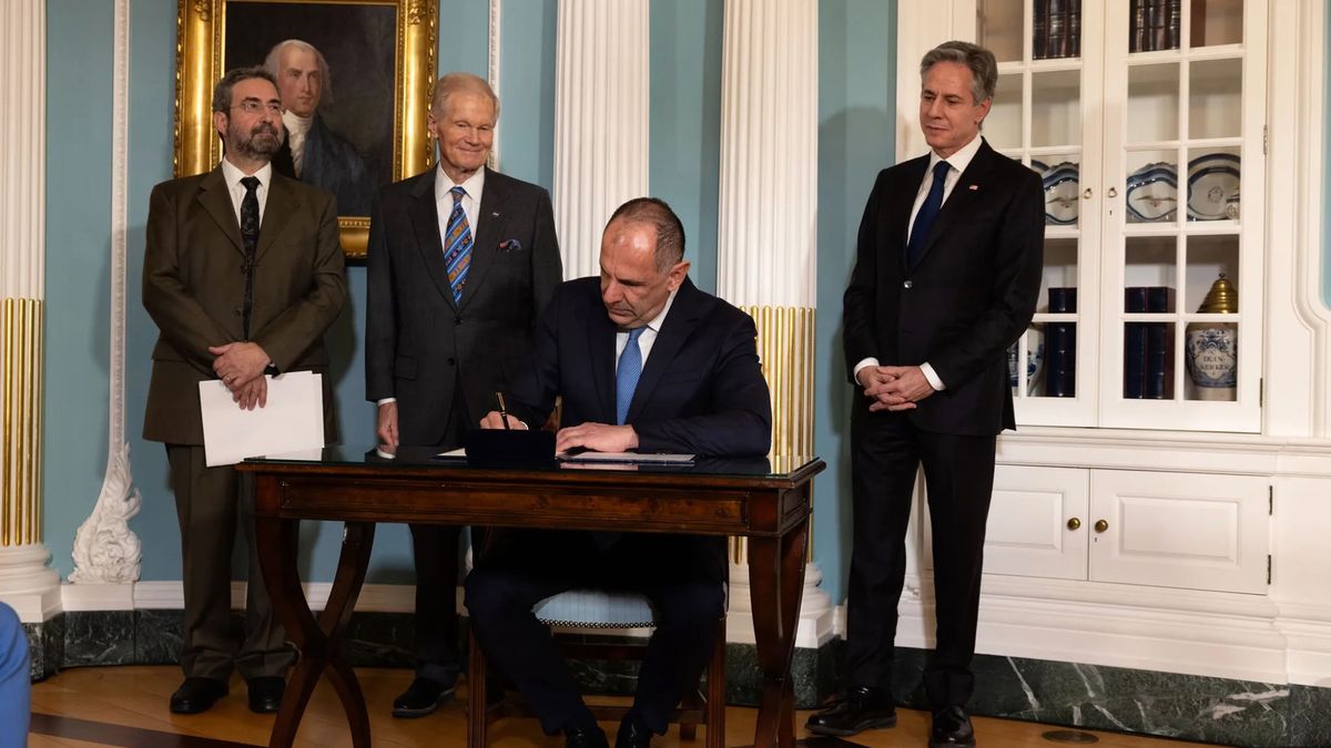 three smiling men in suits stand behind a man who&#039;s sitting at a small wooden table signing a document.