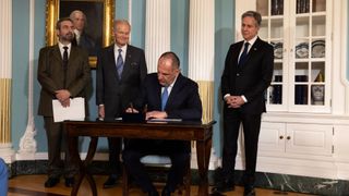 three smiling men in suits stand behind a man who's sitting at a small wooden table signing a document.