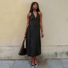woman wears black halterneck dress stood against a wall.