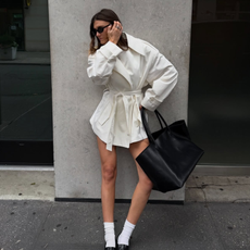Influencer Coco Schiffer wears white coat, black bag, white socks and black loafers standing in front of New York wall.