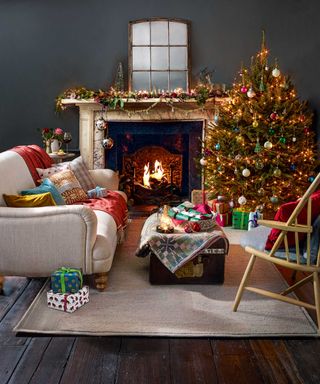 Living room with decorated Christmas tree, neutral sofa and armchair, fireplace and dark painted walls