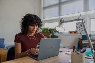 A person with curly hair sits at a desk in a well-lit room, typing on an XPS 13 laptop with notebooks, a lamp, and office supplies nearby.