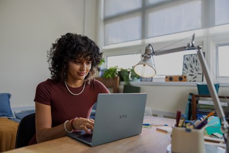 A person with curly hair sits at a desk in a well-lit room, typing on an XPS 13 laptop with notebooks, a lamp, and office supplies nearby.