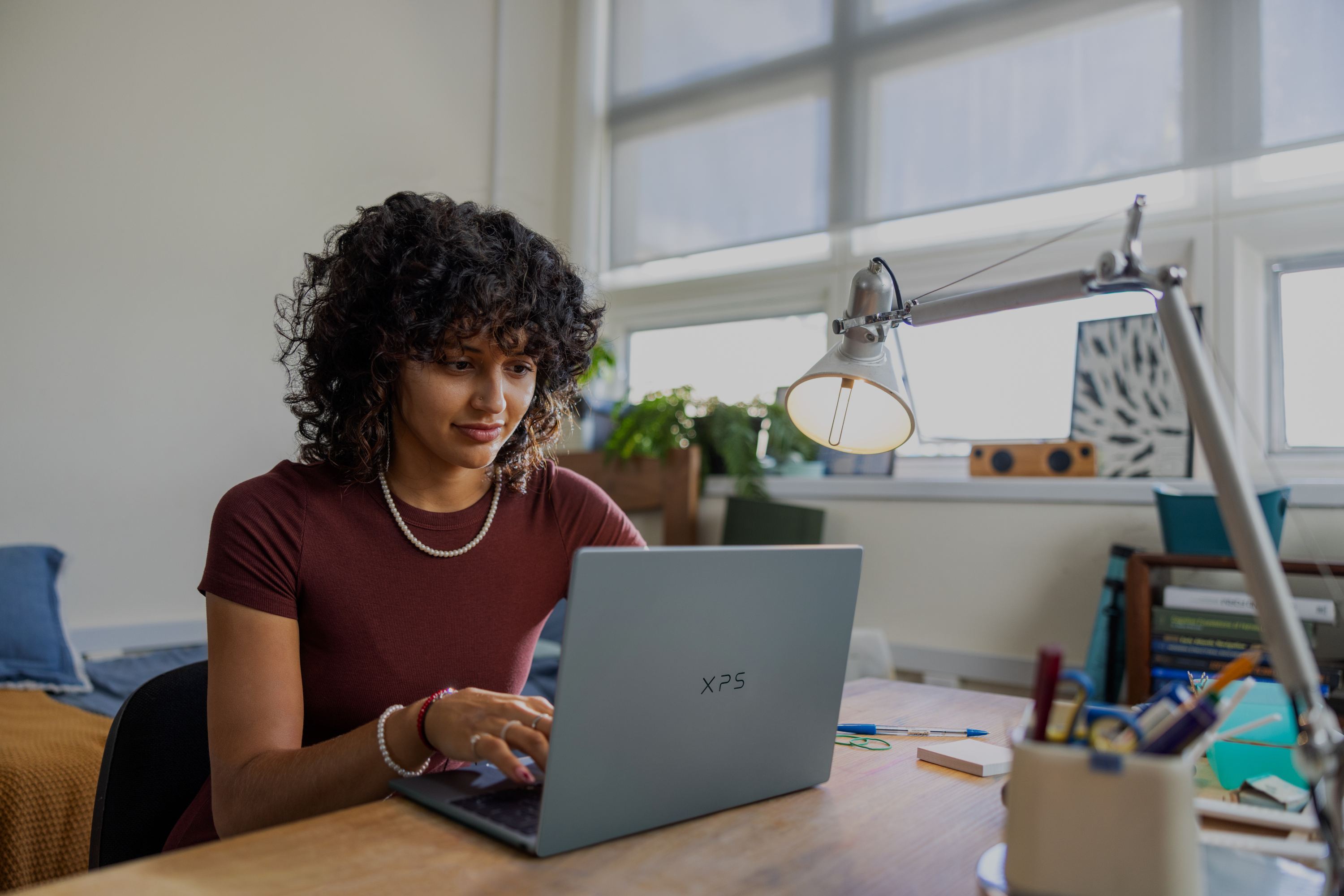 A person with curly hair sits at a desk in a well-lit room, typing on an XPS 13 laptop with notebooks, a lamp, and office supplies nearby.