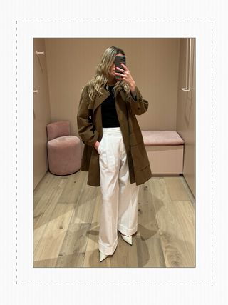 Eliza Huber in the dressing room at Me+Em's new NYC store wearing a brown swing coat with white trousers.