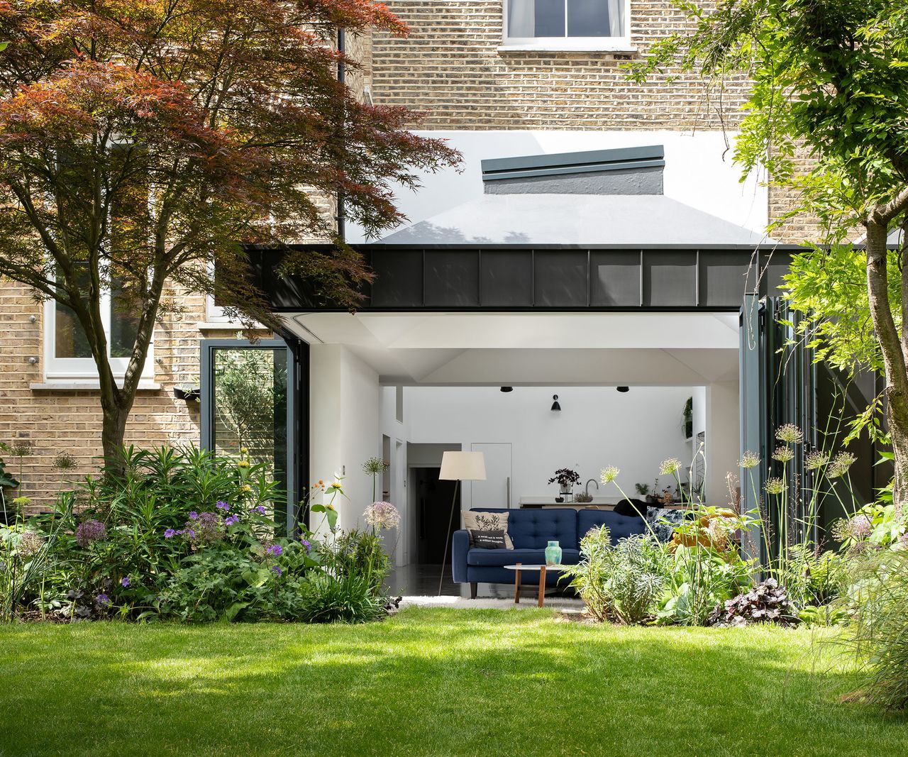 A contemporary kitchen extension with fully open bifold doors and views of a pristine garden lawn and flower beds