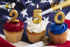 Cupcakes are decorated with red, white, and blue frosting and topped with gold candles that show the number 250. Party favors surround the desserts on a flag backdrop.