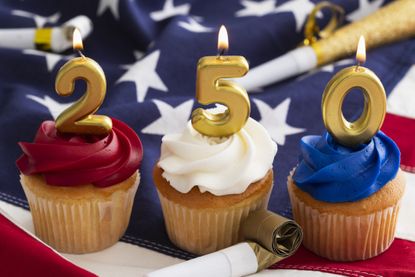 Cupcakes are decorated with red, white, and blue frosting and topped with gold candles that show the number 250. Party favors surround the desserts on a flag backdrop.
