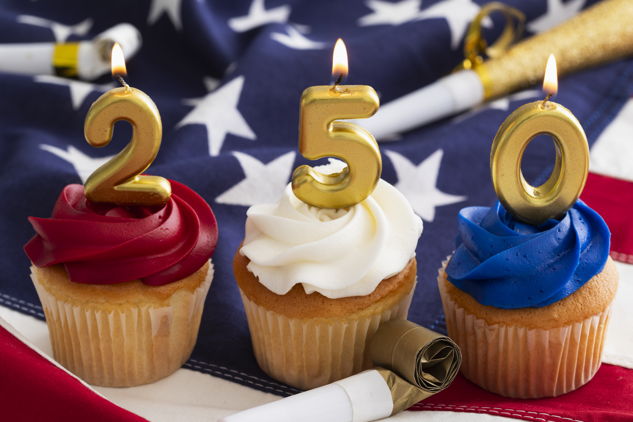 Cupcakes are decorated with red, white, and blue frosting and topped with gold candles that show the number 250. Party favors surround the desserts on a flag backdrop.