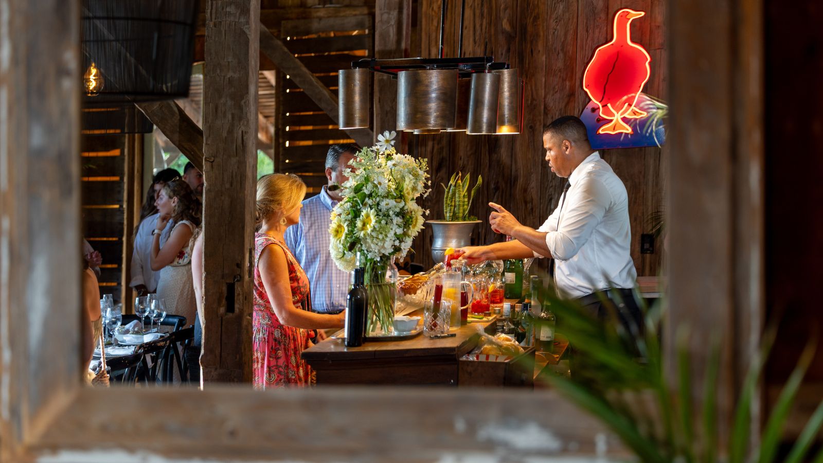 Bar in a rustic barn with a bright red neon light, flowers in vases and a cocktail waiter making drinks