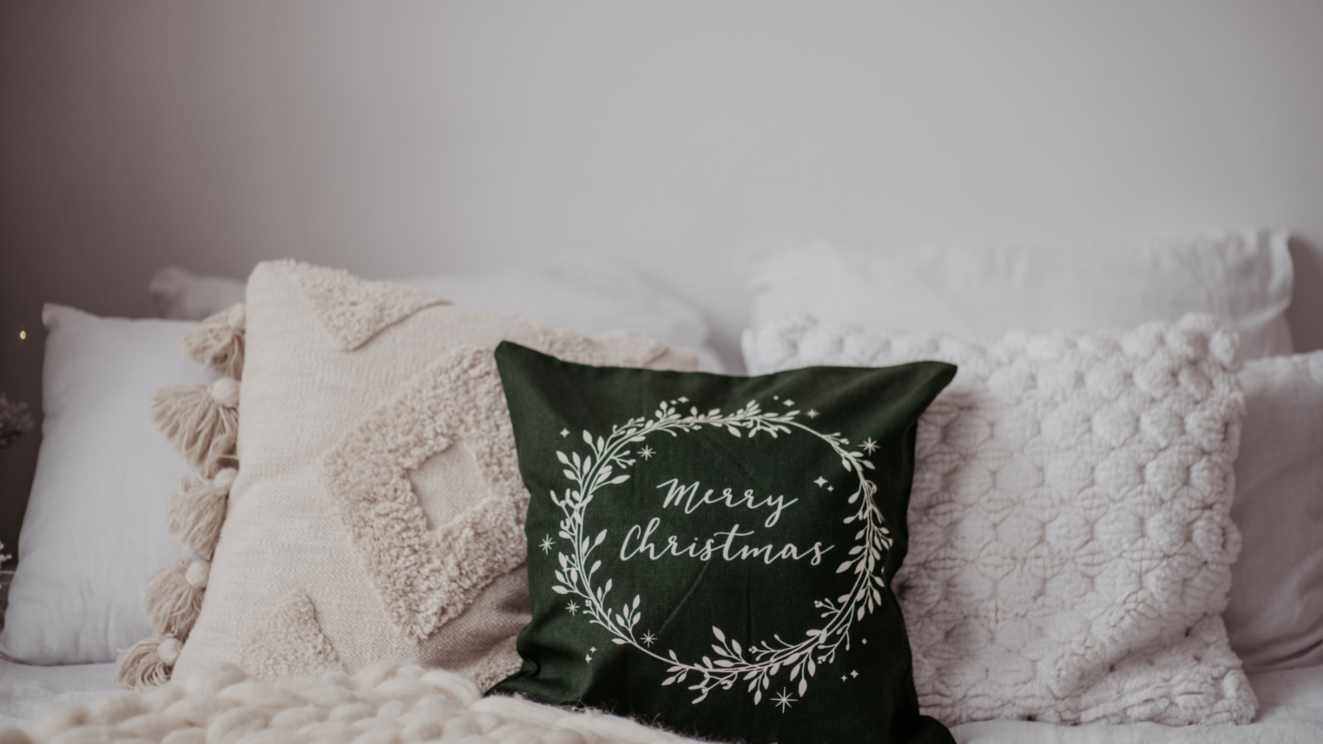A single green Merry Christmas cushion among many white ones in a cozy bedroom space