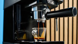 the de'longhi stilosa espresso machine in black, with one portafilter, a dial for espresso and milk steaming, and a plastic tamper, photographed against a blue background