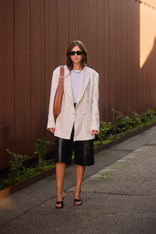 A woman wearing an ivory oversized blazer, a white t-shirt, black heels, a brown shoulder bag, chain necklace, black sunglasses, and black leather bermuda shorts.