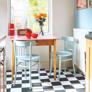 A kitchen dining area with a square table and two light blue chairs on a black and white checkered tiled floor
