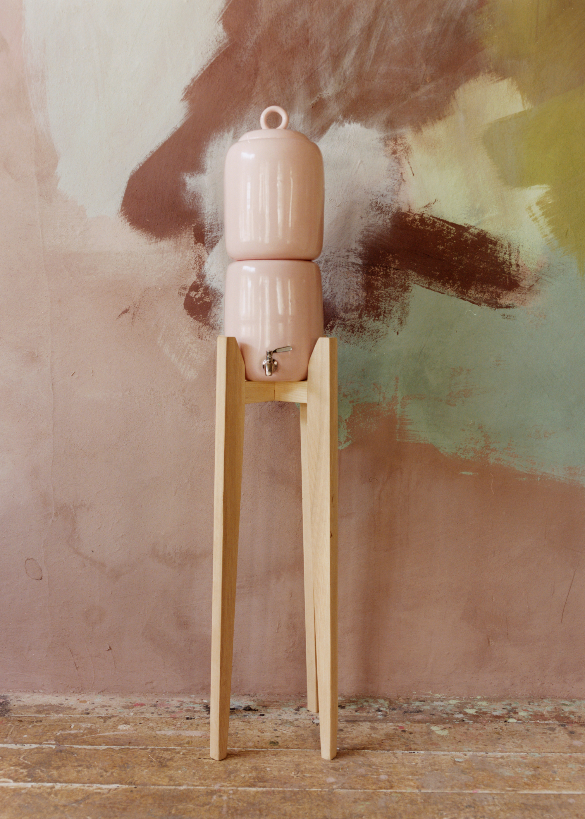 A light beige ceramic water vessel on a wood stand, in front of a patch painted wall