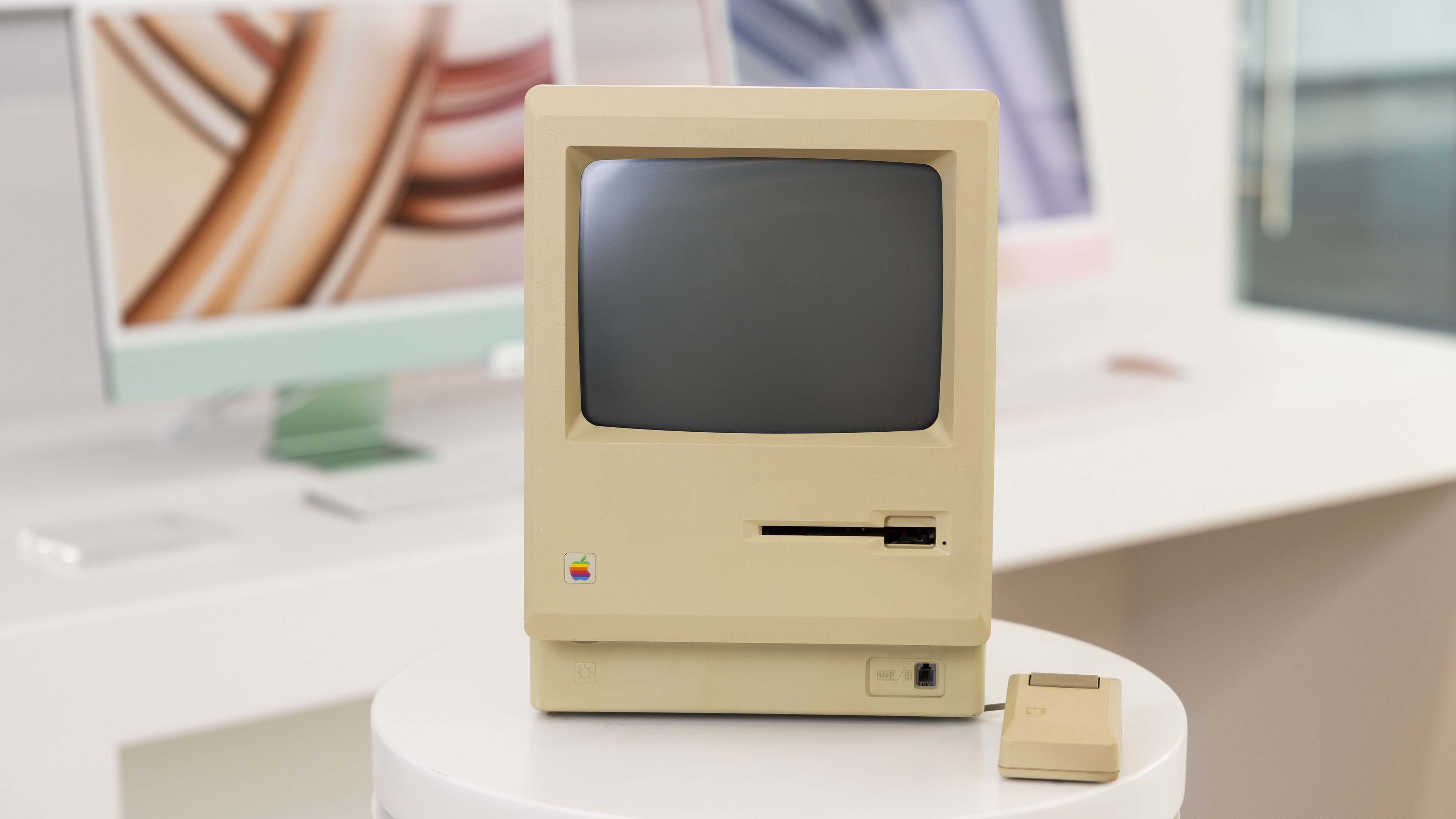 An Apple Macintosh computer with a mouse attached sitting in a room with modern computers behind it.