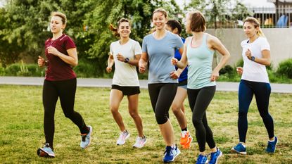 Six women are running together, all of them wearing sports apparel and sneakers. They are running on grass, behind them is a gravel path and some leafy trees. Two of them are having a conversation.