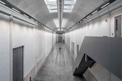 A corridor with tall ceilings, white walls, grey flooring with several grey doors. A view of a behind of a grey steel staircase 