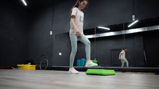 A woman practices step-ups in a gym. There is a low platform in front of her and she is raising her foot to step on it. Behind her is a large mirror.