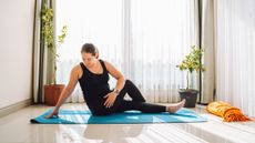 A peaceful looking woman practices yoga on an exercise mat in front of a sunny window. She is on the floor, with one arm behind her on the ground for balance. One of her legs lies straight in front of her, while the other is crossed over it.